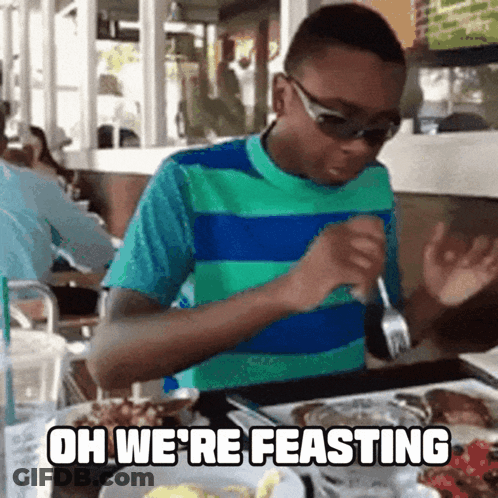 A Boy Is Sitting At A Table With A Plate Of Food And The Words Oh We 'Re Feasting Written Above Him GIF