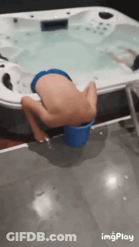 A Boy Is Sitting In A Blue Bucket Near A Hot Tub GIF