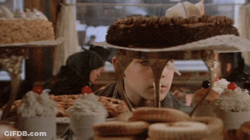 A Boy Is Standing In Front Of A Table Full Of Desserts Including A Chocolate Cake GIF