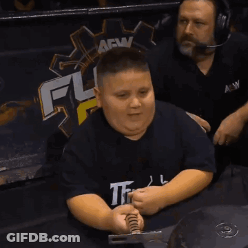 A Boy Wearing A Black Shirt With The Word The On It Sits In Front Of A Sign That Says Aew GIF