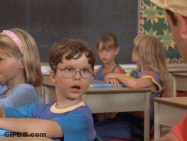 A Boy Wearing Glasses Sits At A Desk In A Classroom With Other Children GIF