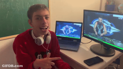 A Boy Wearing Headphones Is Sitting At A Desk In Front Of Two Monitors One Of Which Shows A Dj On It GIF