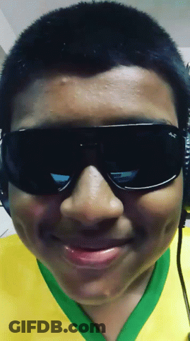 A Boy Wearing Sunglasses And A Yellow And Green Shirt Smiles GIF
