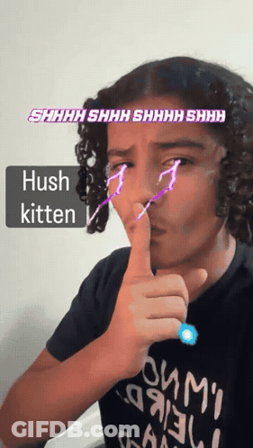 A Boy With Curly Hair Is Making A Hush Kitten Gesture With His Finger GIF