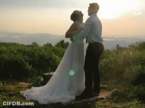 A Bride And Groom Are Dancing In A Field At Sunset . The Bride Is Wearing A Wedding Dress . GIF