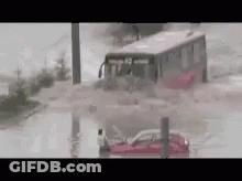 A Bus Is Driving Through A Flooded Street With A Car In The Water . GIF