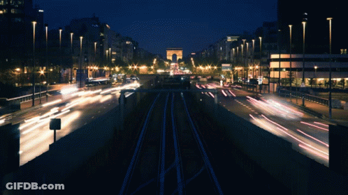 A Busy City Street At Night With A Large Archway In The Background GIF