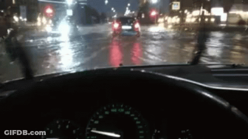 A Car Is Driving Down A Wet Street At Night With A Steering Wheel In The Foreground . GIF
