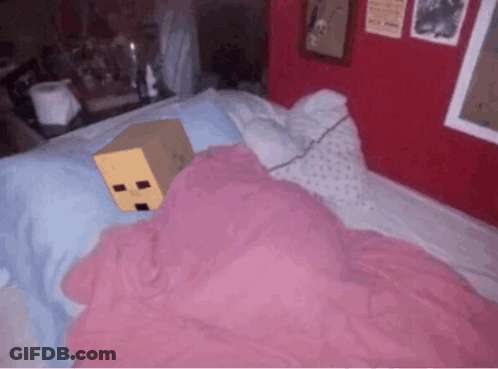 A Cardboard Box With A Face On It Is Laying On A Bed With Pink Sheets GIF