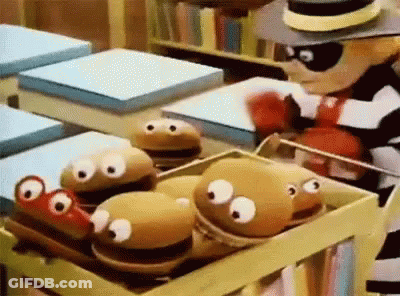 A Cartoon Character Is Standing Next To A Box Of Stuffed Hamburgers With Googly Eyes . GIF
