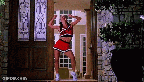 A Cheerleader Is Standing In Front Of A Door With Her Arms In The Air . GIF