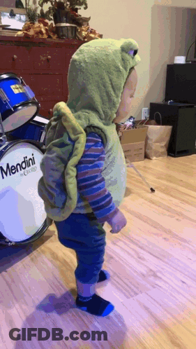 A Child In A Frog Costume Is Standing In Front Of A Drum That Says Mendini GIF