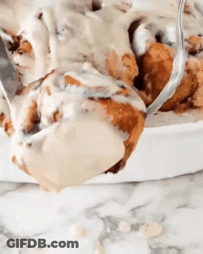 A Close Up Of A Cinnamon Roll With Icing And A Spoon In It . GIF