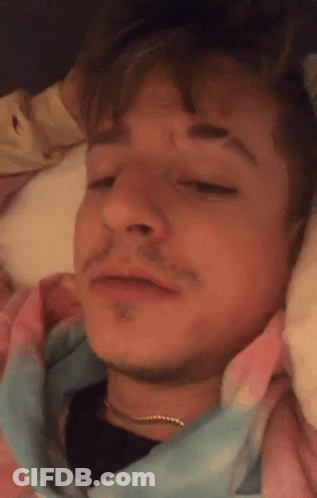 A Close Up Of A Man Laying In Bed With His Eyes Closed And A Tie Dye Shirt On . GIF