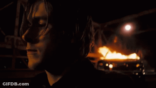 A Close Up Of A Person 'S Face With A Burning Vehicle In The Background GIF
