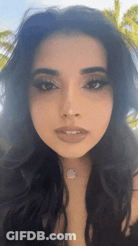 A Close Up Of A Woman 'S Face With A Necklace Around Her Neck And A Palm Tree In The Background GIF