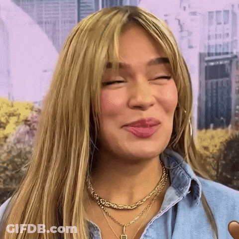 A Close Up Of A Woman Wearing A Necklace And A Denim Shirt Making A Funny Face . GIF
