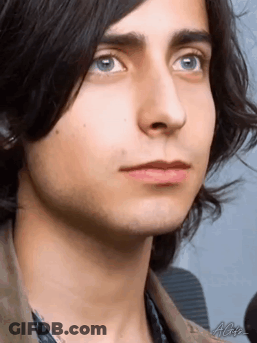A Close Up Of A Young Man 'S Face With Blue Eyes And Long Black Hair . GIF