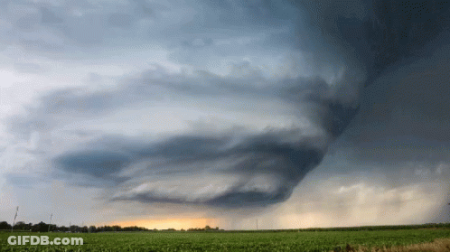 A Cloudy Sky Over A Field With A Few Trees In The Distance GIF