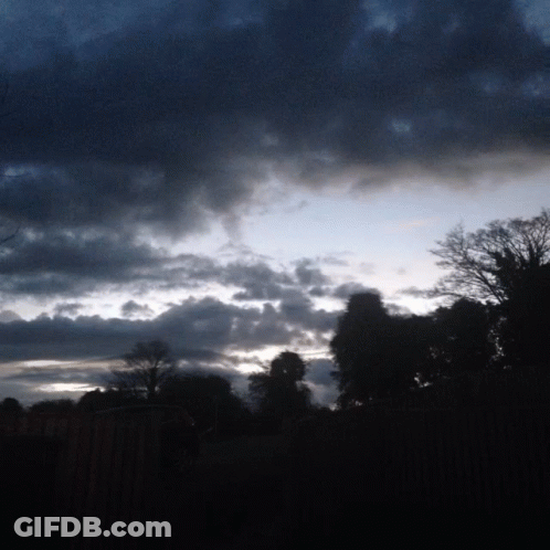 A Cloudy Sky With Trees In The Foreground And A Fence In The Background GIF