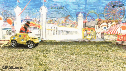 A Clown Is Driving A Toy Car In Front Of A Cyclone Ride GIF