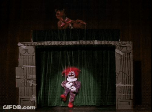 A Clown Puppet Is Being Held Up By A String On A Stage GIF