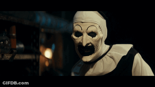 A Clown Wearing A White Mask With A Black Mouth Is Standing In A Dark Room GIF