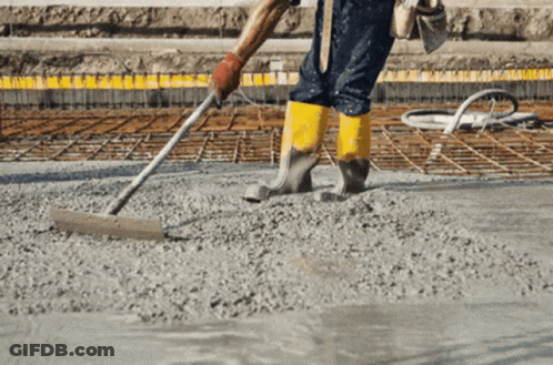 A Construction Worker Wearing Yellow Boots Spreads Concrete With A Rake GIF