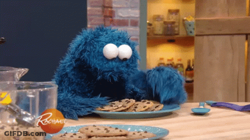 A Cookie Monster Is Sitting At A Table With A Plate Of Chocolate Chip Cookies GIF