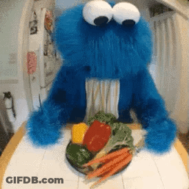 A Cookie Monster Is Sitting At A Table With A Plate Of Vegetables In Front Of Him GIF