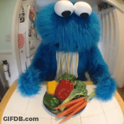 A Cookie Monster Is Sitting At A Table With A Plate Of Vegetables In His Mouth GIF