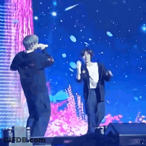 A Couple Of People Standing On A Stage Holding Microphones GIF