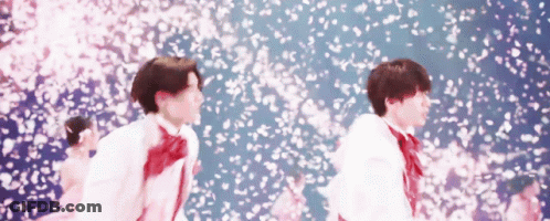 A Couple Of Young Men Are Standing In Front Of A Wall Of Cherry Blossoms . GIF