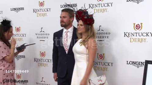 A Couple Standing In Front Of A Longines Kentucky Derby Sign GIF