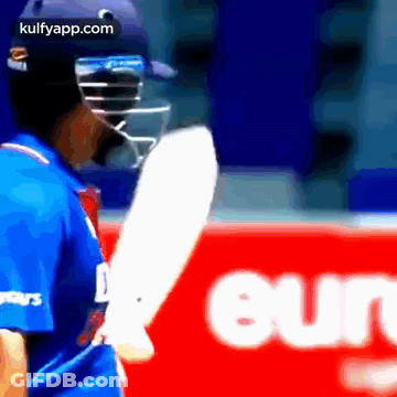 A Cricket Player Is Wearing A Helmet And Holding A Bat In Front Of A Red Sign . GIF