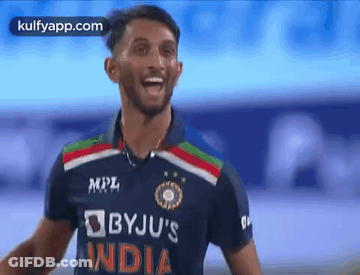 A Cricket Player Wearing A Blue And Red Jersey Is Standing On A Field And Smiling . GIF