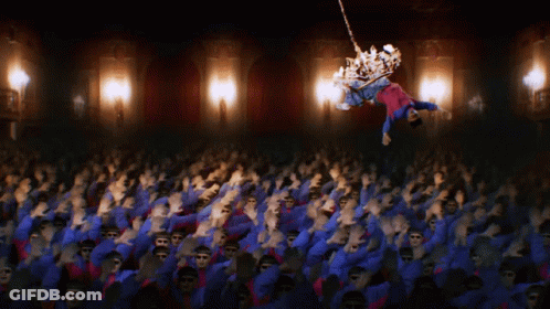 A Crowd Of People Are Dancing In A Dark Room With A Chandelier In The Background GIF