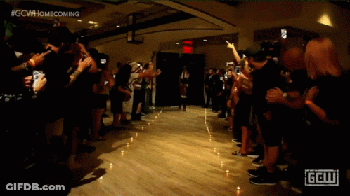 A Crowd Of People Are Gathered In A Dark Room With A Sign That Says Gcw Homecoming GIF
