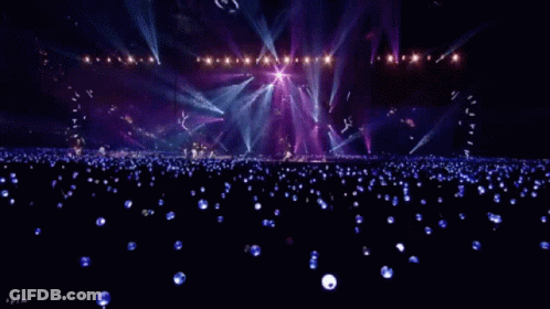 A Crowd Of People Are Sitting In A Stadium Watching A Concert With Purple Lights . GIF