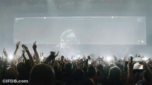 A Crowd Of People At A Concert With Their Hands In The Air In Front Of A Large Screen With A Skull On It GIF