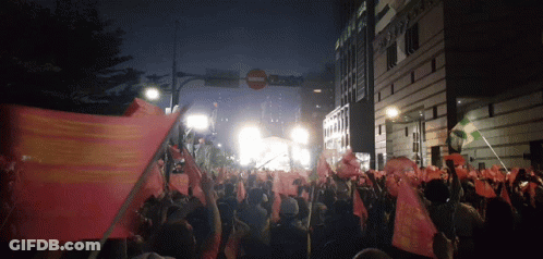 A Crowd Of People Holding Up Flags In Front Of A Building That Says ' Sydney Bank ' On It GIF