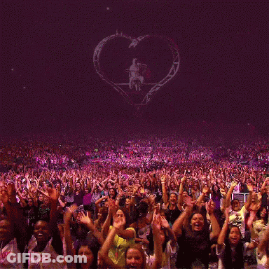 A Crowd Of People With Their Hands In The Air At A Concert With A Heart In The Background GIF