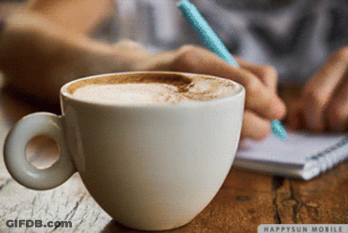 A Cup Of Coffee Sits On A Table Next To A Person Writing On A Notebook GIF