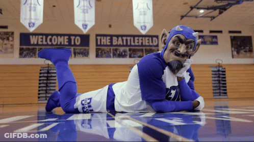 A Devil Mascot Is Laying On The Floor In Front Of A Sign That Says Welcome To The Battlefield GIF