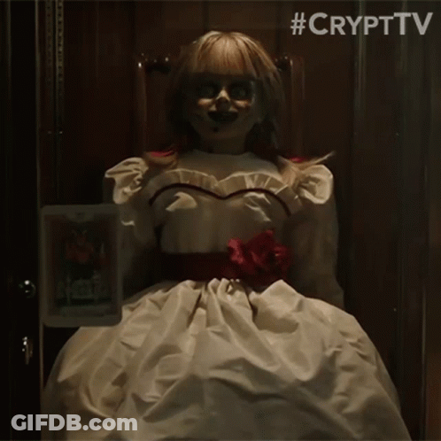 A Doll In A White Dress Is Sitting In A Chair Holding A Picture Of Annabelle . GIF