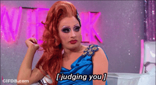 A Drag Queen In A Blue Dress Is Sitting In Front Of A Pink Sign That Says Week GIF