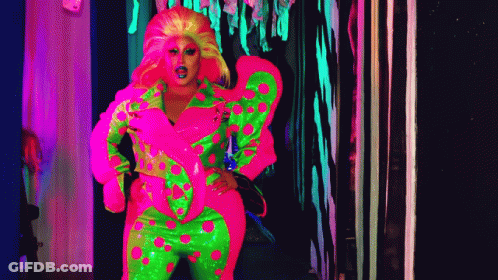 A Drag Queen In A Neon Pink And Green Outfit Is Standing In A Dark Hallway . GIF