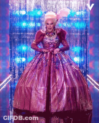 A Drag Queen In A Purple And Gold Dress With A V On The Bottom GIF