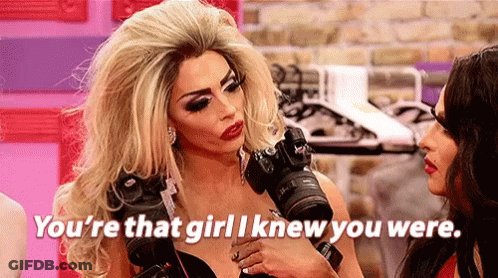 A Drag Queen Is Holding A Camera In Her Hand And Talking To Another Woman . GIF
