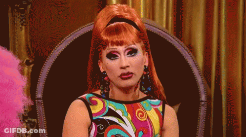 A Drag Queen Is Sitting In A Chair Wearing A Colorful Dress GIF